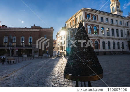Cobblestone square in Riga's old town featuring a cone shaped Christmas Tree sculpture, Riga Town Hall with clock tower, and historic architecture. 130807408