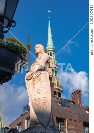 A knight statue with a shield stands before St. Peter's Church spire in Riga, Latvia, with a blue sky, clouds, and yellow flowers in the foreground. 130807412