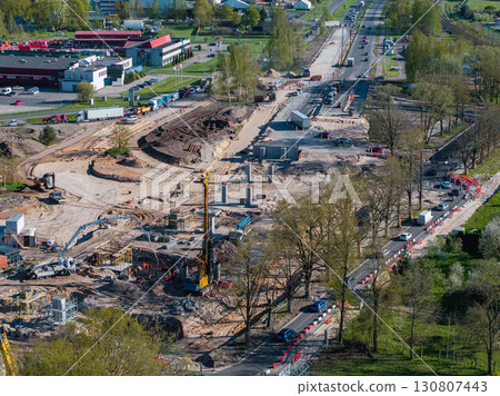 Active construction site in Riga, Latvia, showing cranes, excavators, foundations, structural columns, nearby roads, green spaces, and buildings. 130807443