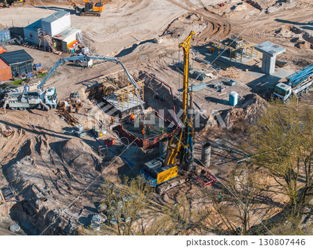 Active construction site with a yellow drilling rig, workers, machinery, and materials. Trees and scattered equipment frame the infrastructure project. Active construction site with a yellow drilling rig, workers, machinery, and materials. Trees and scattered equipment frame the infrastructure project. 130807446