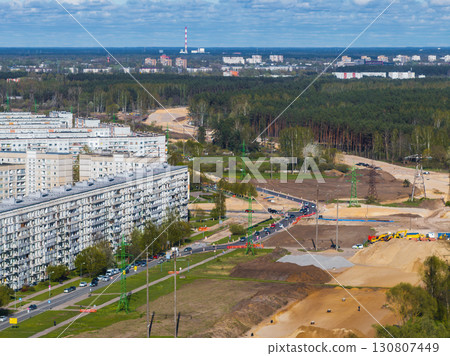 View of a construction site in Riga, Latvia, with sand piles, equipment, roadwork, a residential block, tree lined street, forest, and a red and white chimney. 130807449