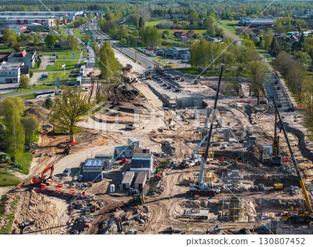Active construction site in Riga, Latvia, with cranes, excavators, workers, concrete structures, green spaces, residential buildings, and light traffic nearby. 130807452
