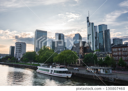 Skyline of Frankfurt, Germany, featuring modern skyscrapers like Main Tower, a historic church, the River Main, a docked boat, and green trees. 130807468