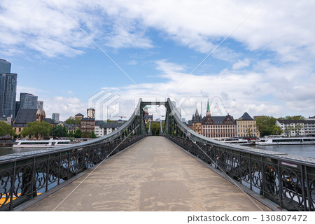 The Eiserner Steg bridge spans the Main River in Frankfurt, Germany, with ornate ironwork, city skyscrapers, a church spire, and tree lined riverbanks. The Eiserner Steg bridge spans the Main River in Frankfurt, Germany, with ornate ironwork, city skyscrapers, a church spire, and tree lined riverbanks. 130807472