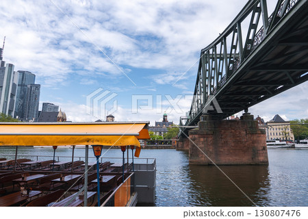 The Eiserner Steg bridge spans the Main River in Frankfurt, Germany, with steel and stone details, a docked yellow roofed boat, and city skyscrapers. The Eiserner Steg bridge spans the Main River in Frankfurt, Germany, with steel and stone details, a docked yellow roofed boat, and city skyscrapers. 130807476