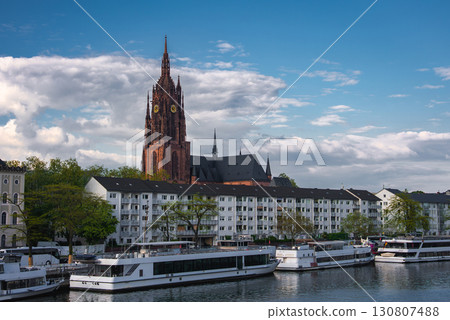 The Frankfurt Cathedral's Gothic spire rises against a partly cloudy sky, with the Main River, riverboats, residential buildings, and greenery in view. The Frankfurt Cathedral's Gothic spire rises against a partly cloudy sky, with the Main River, riverboats, residential buildings, and greenery in view. 130807488