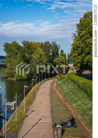 A paved riverside pathway in Frankfurt, Germany, bordered by greenery and a calm river. A motorcycle is parked near the path, with a skyscraper visible. 130807515
