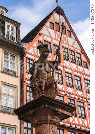A detailed statue of a warrior holding a spear stands on an ornate pedestal, with a red and white half timbered building and modern architecture in Frankfurt. A detailed statue of a warrior holding a spear stands on an ornate pedestal, with a red and white half timbered building and modern architecture in Frankfurt. 130807570