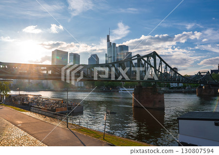 The Frankfurt skyline features modern skyscrapers like Commerzbank Tower, the Eiserner Steg bridge over the Main River, and a lively riverside promenade. 130807594