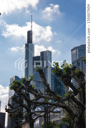 The Frankfurt skyline in Germany with the Commerzbank Tower, modern high rises, pruned trees in the foreground, and a bright blue sky with clouds. The Frankfurt skyline in Germany with the Commerzbank Tower, modern high rises, pruned trees in the foreground, and a bright blue sky with clouds. 130807604