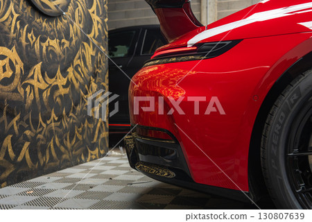 Rear view of a red 2025 Porsche 911 GT3 922 with sleek curves and taillight, parked indoors on a checkered floor with graffiti style wall behind. Rear view of a red 2025 Porsche 911 GT3 922 with sleek curves and taillight, parked indoors on a checkered floor with graffiti style wall behind. 130807639