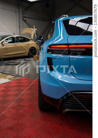 Rear view of a blue Porsche Macan on a red floor mat in a garage. A beige car with its hood open and tools are visible in the industrial space. 130807679