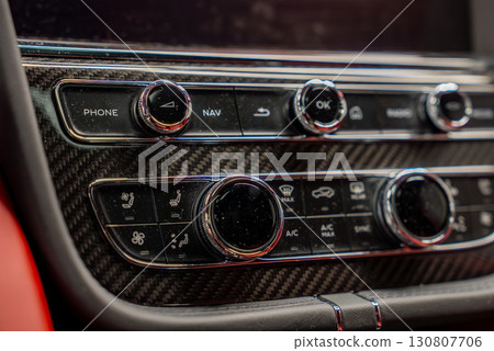 Detailed view of a Bentley Bentayga control panel featuring carbon fiber trim, chrome knobs, and red leather accents in the background. 130807706