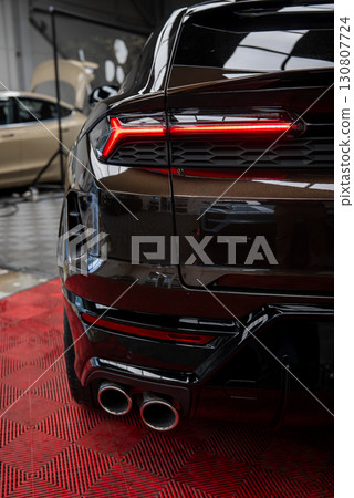 Rear view of a black Lamborghini Urus with dual exhaust pipes and red taillights, parked indoors on a red floor with another luxury car in the background. 130807724
