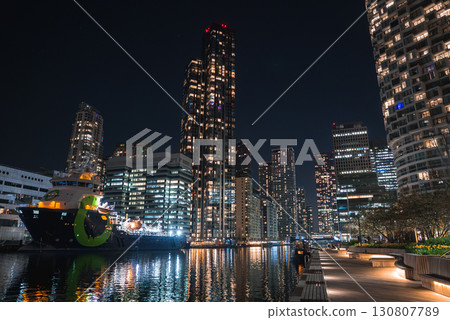 Canary Wharf's illuminated skyscrapers at night, a green hulled docked ship, calm canal waters, a lit boardwalk, benches, and landscaped greenery. 130807789
