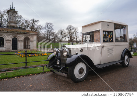 A vintage white and black ice cream van is parked on a paved path near a neoclassical building with arched doorways, surrounded by grass and flowerbeds. A vintage white and black ice cream van is parked on a paved path near a neoclassical building with arched doorways, surrounded by grass and flowerbeds. 130807794