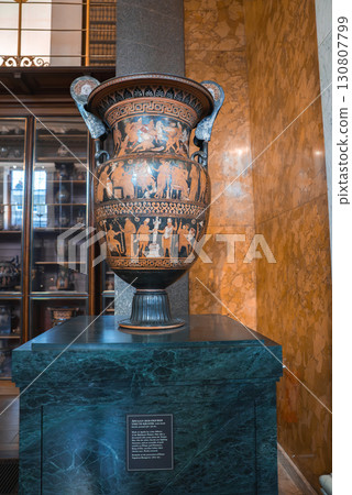 Large decorated vase with red figure artwork on a green marble pedestal, displayed in a museum with golden walls and a glass cabinet of artifacts. 130807799
