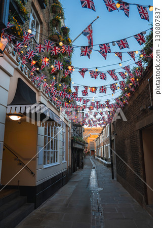 A narrow London street adorned with Union Jack flags, brick and stucco buildings, flower boxes, warm lights, and a cobblestone path leading into the distance. A narrow London street adorned with Union Jack flags, brick and stucco buildings, flower boxes, warm lights, and a cobblestone path leading into the distance. 130807837