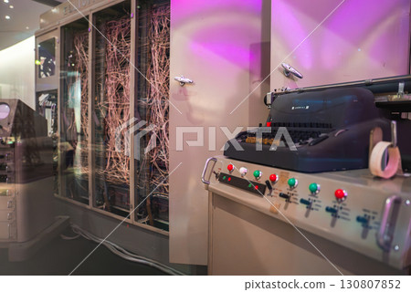 A retro computing setup with a typewriter style input, colorful control panel, and exposed wiring in a softly lit exhibit, possibly in London. A retro computing setup with a typewriter style input, colorful control panel, and exposed wiring in a softly lit exhibit, possibly in London. 130807852