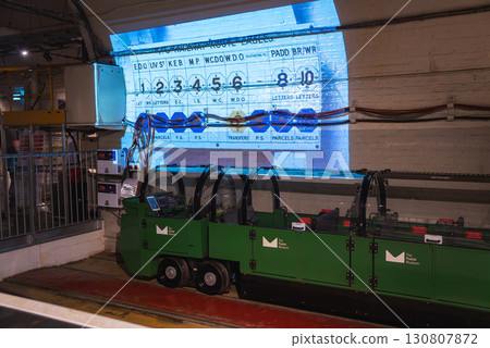 A green train with transparent covers on the Mail Rail tracks in London. A projection shows a vintage postal sorting board in an industrial setting. 130807872