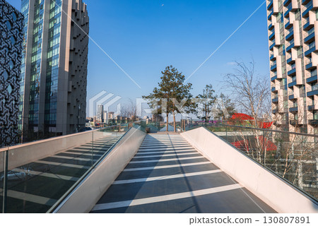 A glass railed walkway leads to trees and an open area, surrounded by high rise buildings with unique facades under a clear blue sky in London. A glass railed walkway leads to trees and an open area, surrounded by high rise buildings with unique facades under a clear blue sky in London. 130807891