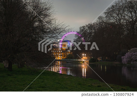 The London Eye glows in purple hues, partially obscured by trees. A calm body of water reflects the Ferris wheel and nearby buildings in a tranquil park setting. The London Eye glows in purple hues, partially obscured by trees. A calm body of water reflects the Ferris wheel and nearby buildings in a tranquil park setting. 130807924