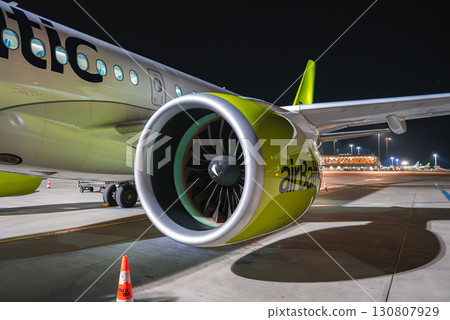 A parked airBaltic aircraft on a tarmac at night, featuring a bright green engine, visible branding, a lit terminal, and faintly seen nearby planes. A parked airBaltic aircraft on a tarmac at night, featuring a bright green engine, visible branding, a lit terminal, and faintly seen nearby planes. 130807929
