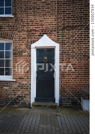Classic brick facade of a London style townhouse featuring a green door with a brass lion head knocker, a white framed window, and gray brick paving. Classic brick facade of a London style townhouse featuring a green door with a brass lion head knocker, a white framed window, and gray brick paving. 130807934
