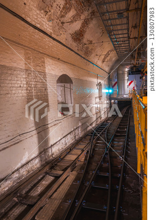 Aged brick walls and dual railway tracks curve into the distance in a London underground tunnel. Overhead pipes and a metal walkway add industrial detail. 130807983