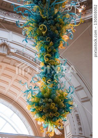 Intricate glass sculpture in blue, green, and yellow suspended in an ornate interior with arched window and coffered ceiling at a historic museum. Intricate glass sculpture in blue, green, and yellow suspended in an ornate interior with arched window and coffered ceiling at a historic museum. 130808002