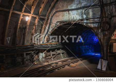 Aged underground tunnel in London with exposed concrete walls, structural reinforcements, and tracks leading into a dimly lit section with blue light. 130808004