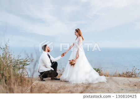 Groom kneeling in front of bride by the sea 130810425