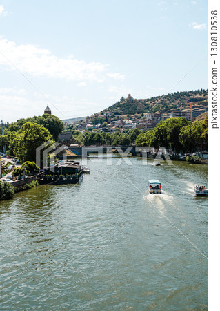 Serene river view with boats passing by in a vibrant city near the hillside during a sunny day Serene river view with boats passing by in a vibrant city near the hillside during a sunny day 130810538