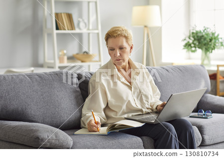Mature woman is focused on work, taking notes in notebook while using her laptop. Mature woman is focused on work, taking notes in notebook while using her laptop. 130810734