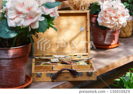Old wooden paint box with brushes surrounded by blooming flowers in pots on sunny wooden table Old wooden paint box with brushes surrounded by blooming flowers in pots on sunny wooden table 130810829