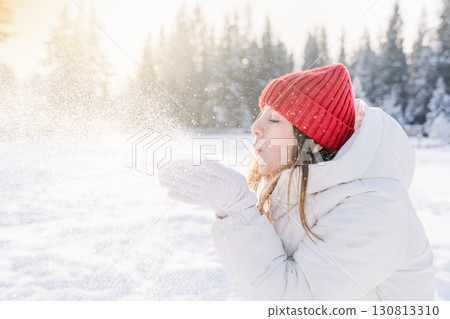 Young woman in winter attire playfully blowing snowflakes in a sunlit snowy landscape surrounded by evergreens Young woman in winter attire playfully blowing snowflakes in a sunlit snowy landscape surrounded by evergreens 130813310
