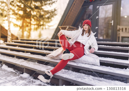 n a serene winter landscape, a woman sits comfortably on wooden stairs adorned with snow. She wears a stylish red outfit and a warm beanie, stretching her leg while enjoying the peaceful atmosphere. 130813316