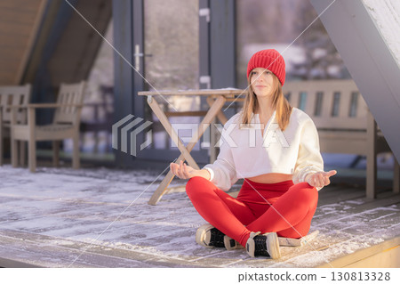 A young thirty-year-old woman in a red riding hood meditates sitting in a lotus position near a house in nature in winter, where she came to relax for the winter holidays and Christmas. 130813328