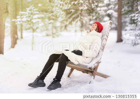 A woman in a white down jacket sits reclining on a wooden armchair in a snowy forest. Relaxing in nature after the Christmas holidays 130813355