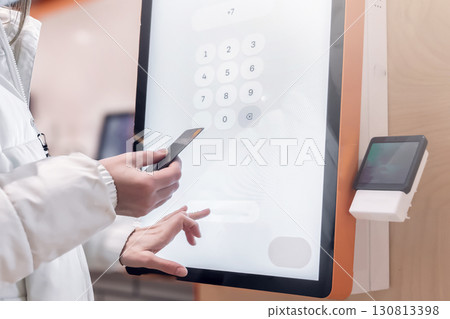 A user engages with a digital kiosk that facilitates seamless and contactless payment processing for transactions. The woman pays through the payment terminal with a credit card. The touch screen 130813398