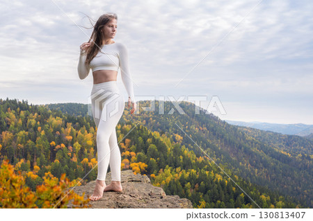 A beautiful young woman in white leggings poses on top of a mountain. Outdoor health and sports. Athletic woman practicing yoga on a mountain peak surrounded by autumn foliage under a cloudy sky 130813407
