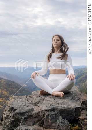 Meditating woman in lotus position on mountain cliff during autumn surrounded by nature. A woman in white relaxes on a mountaintop - outdoor sports in nature 130813408