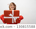 A cheerful woman in festive attire, wearing a jolly Santa hat and red gloves, holds a beautifully wrapped gift, embodying the joyful holiday spirit of giving that unites everyone in celebration 130815588