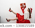 A joyful woman in a vibrant red dress and playful Santa hat holds a wrapped gift, embodying the true spirit of Christmas cheer and spreading warmth and happiness this festive season 130815589