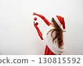 A cheerful woman in a festive Santa hat and elegant red outfit balances a shiny Christmas ornament on her finger, capturing the joyous spirit and warmth of the holiday season around her 130815592