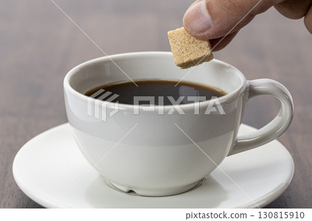 Hand holding organic sugar cube above hot black coffee in white ceramic cup on saucer, preparing to sweeten drink, morning beverage concept, close up, calm mood 130815910