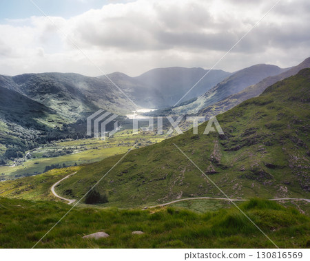 If you look at the landscape, you can see green hills under a grey sky. A curving road goes through the green valley of Gap of Dunloe in County Kerry, Ireland. 130816569