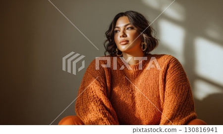 A beautiful plus-size woman in an orange knit sweater sits peacefully in soft autumn sunlight 130816941