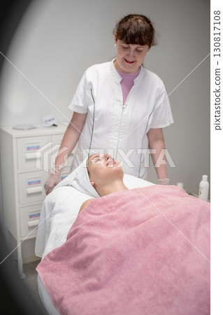 Beautician preparing female client for cosmetic facial treatment in professional beauty salon 130817108