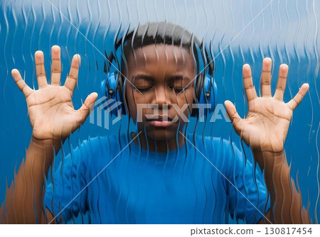 Autistic black child boy with blue noise cancelling headphones pressing hands to textured glass, eyes closed, concept of autism, sensory overload and isolation 130817454
